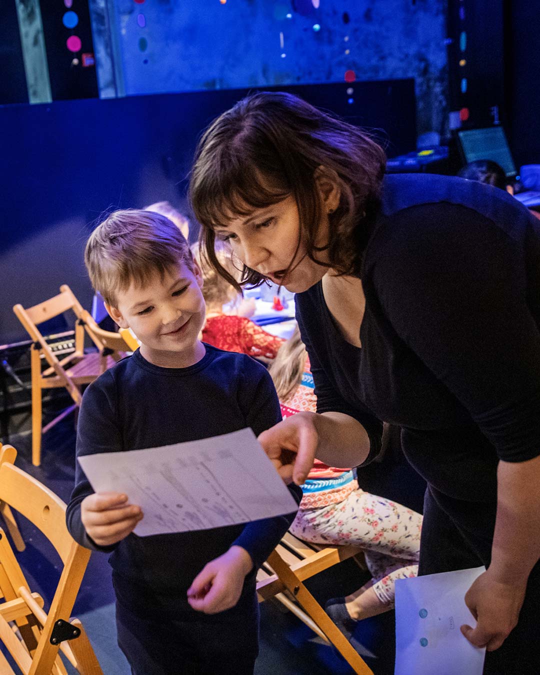 Barn sitter vid bord och ritar. Ett barn visar sin teckning åt en pedagog.