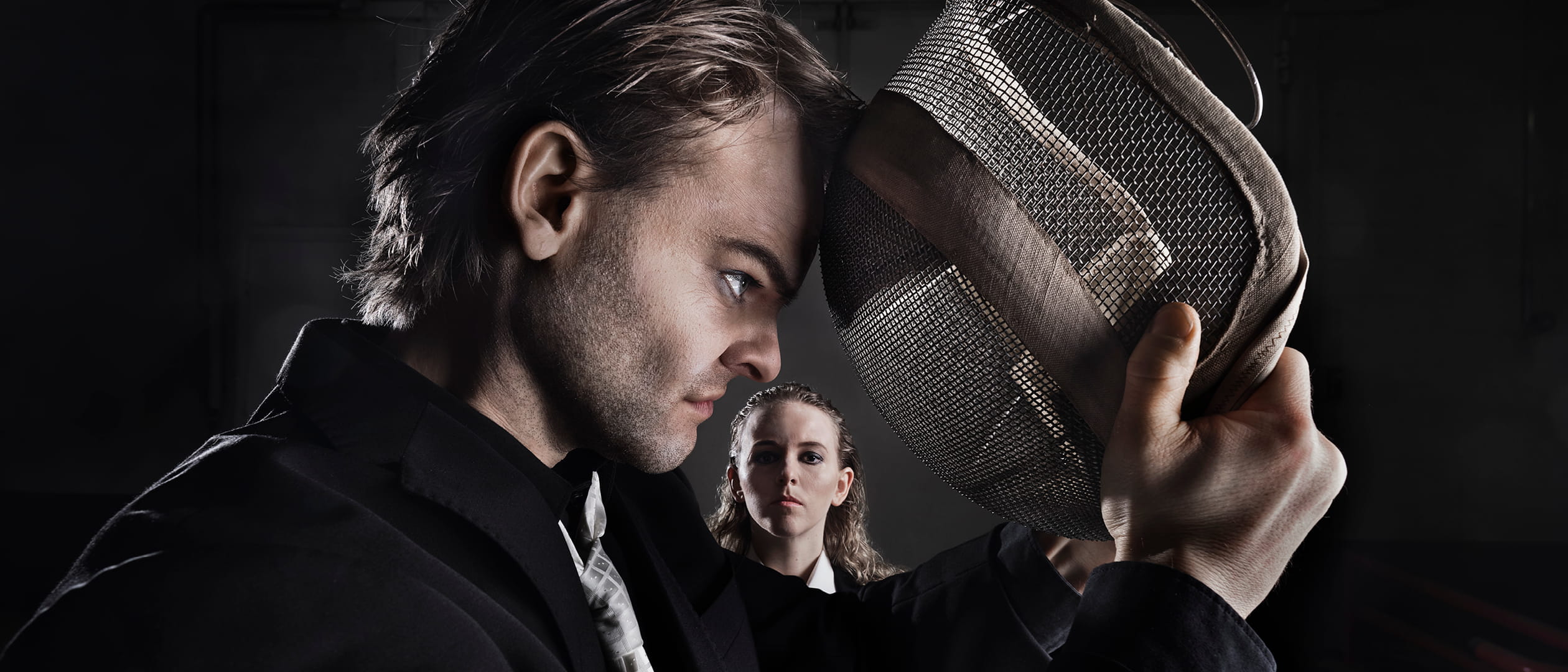 Hamlet nojaa päänsä miekkailunaamiota vasten. Nainen näkyy taustalla.